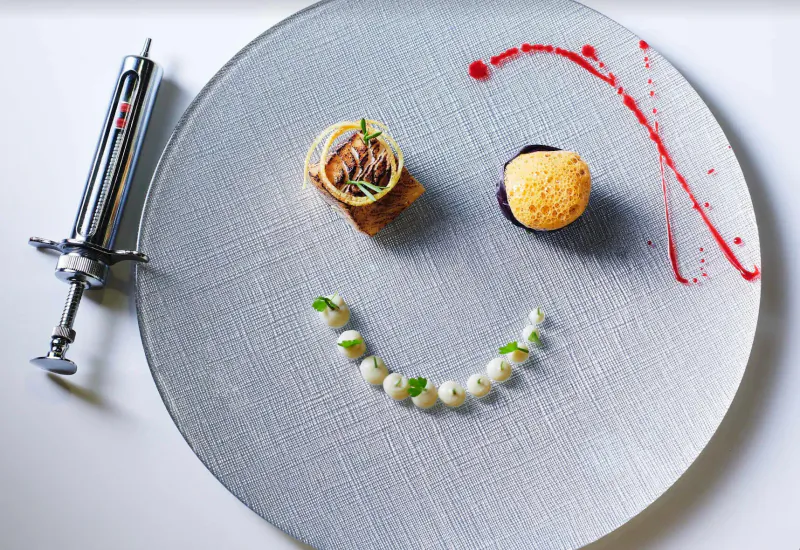 Playful smiley face plate: meat cube eyes, meringue nose, peas mouth, sauce streaks, syringe beside.