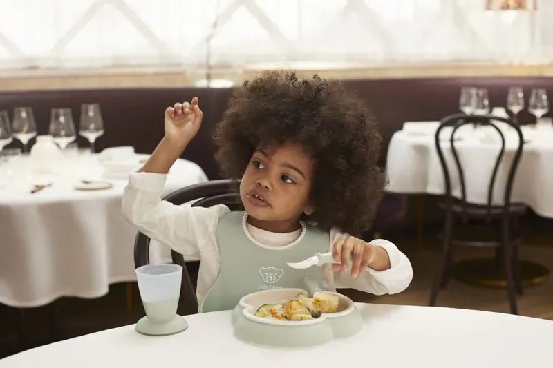 Black toddler girl in green bib eating yellow food from matching bowl and spoon at elegant restaurant table