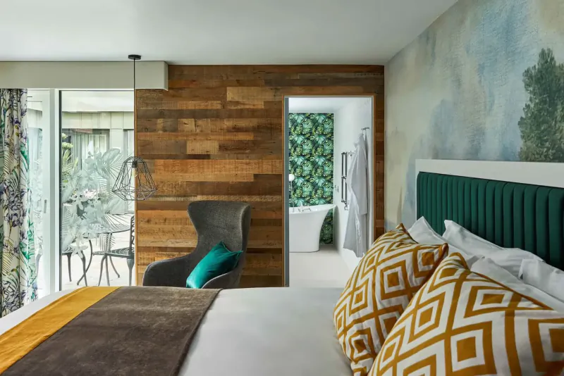 Hotel Indigo Bath hotel room with green velvet headboard bed, pillows, turquoise armchair, wood wall, mural, and bathroom.