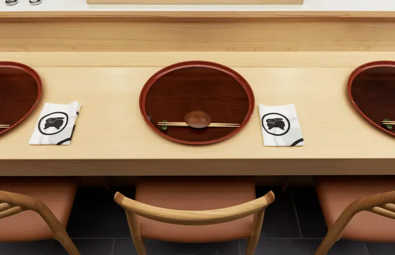 Sushi counter with three red-lacquered plates holding fish on rice, napkins with black logo, wooden stools below.