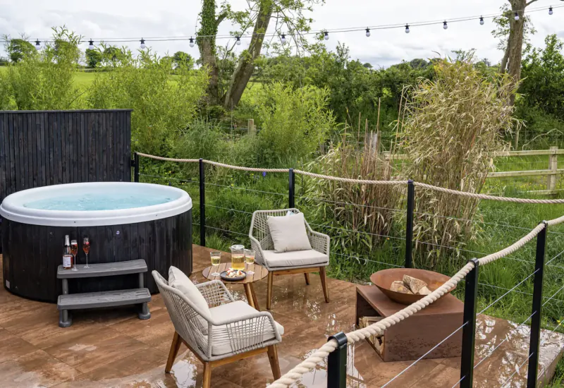 Luxury glamping pod deck with bubbling hot tub, cushioned chairs, wine glasses, fire pit, and rural Welsh countryside view.