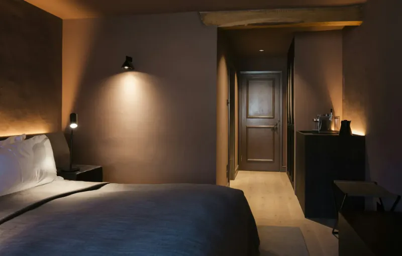 Cozy hotel bedroom with gray bed, warm wall lights, wooden door, and minimalist decor in dim lighting.