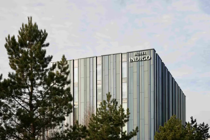 Hotel Indigo Coventry, modern striped high-rise building with 'Hotel Indigo' sign, framed by pine trees under cloudy sky