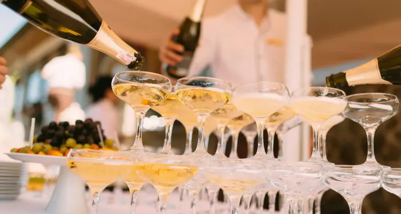 Staff pouring champagne from bottles into a tall stack of glasses at Le Jardin Privé event, with fruit platter nearby.