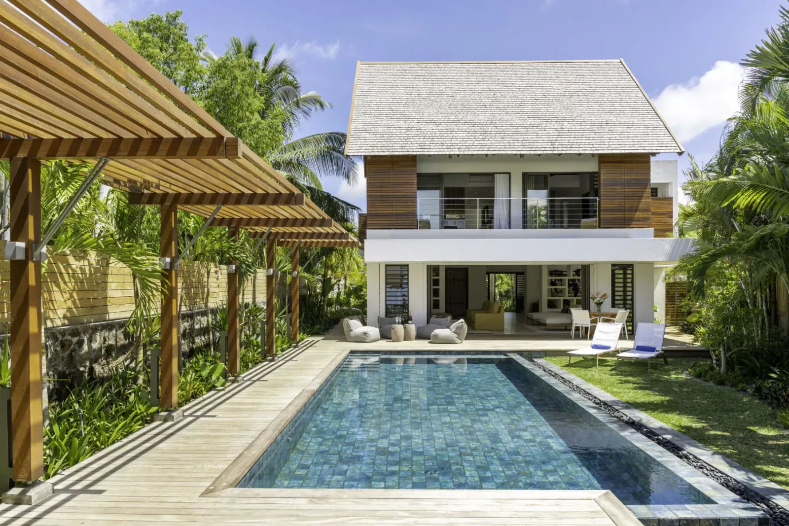 Modern tropical villa with wooden pergola, infinity pool, lounge chairs, and lush palm gardens in Mauritius.