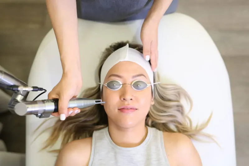 Therapist using laser device on client's face during treatment, both wearing eye protection on white bed.