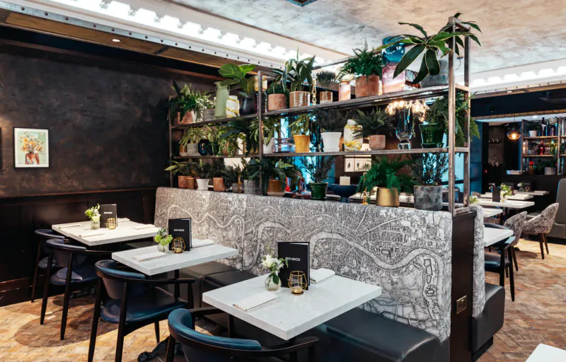 Six by Nico Canary Wharf restaurant interior with marble-patterned dividers, potted plants, set tables, and Ancient Rome decor.