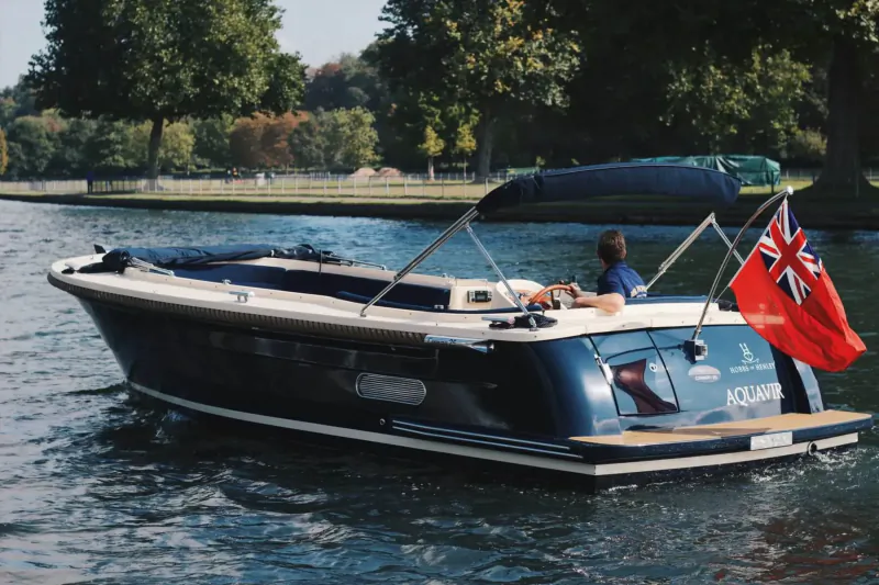 Rear view of Hobbs luxury motorboat with Union Jack flag, steered by person on river amid trees