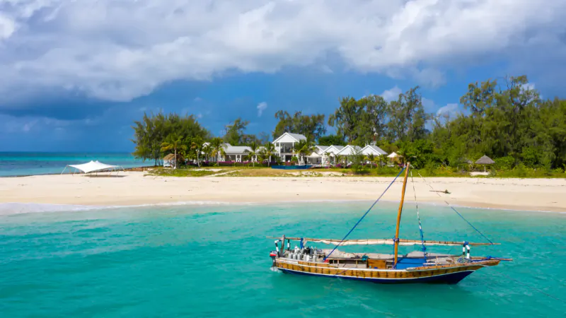 Traditional dhow sailboat anchored in turquoise lagoon off tropical island resort with white buildings and beach.
