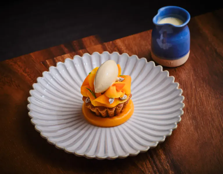 Orange mango tart topped with cream on white scalloped plate, blue milk jug beside, on wooden table