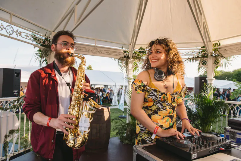 Bearded man with sax and curly-haired woman DJing at outdoor festival under tent, Taste of London.
