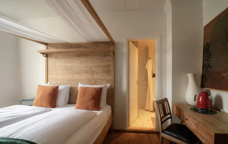 Cozy hotel room with wooden canopy bed, orange pillows, open bathroom door, desk, vase, and red kettle