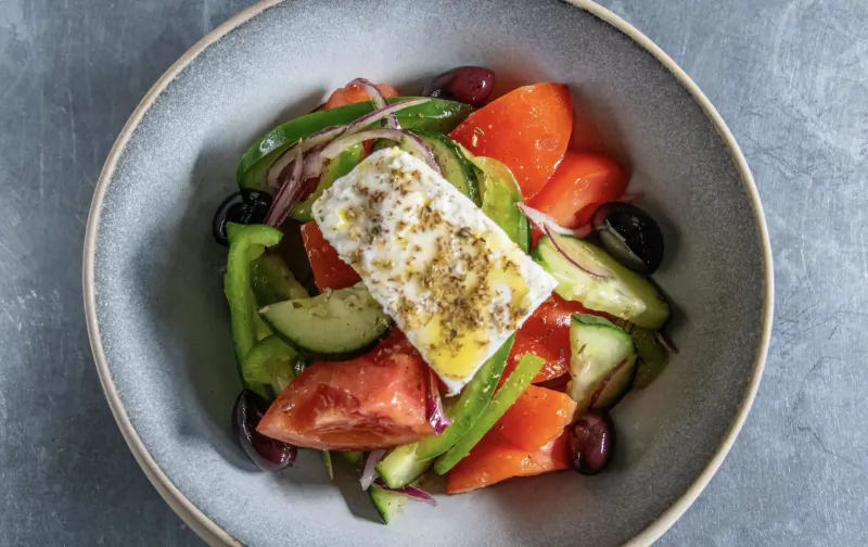 Greek salad in gray bowl with feta cheese, tomatoes, cucumbers, peppers, olives, and onions on gray surface