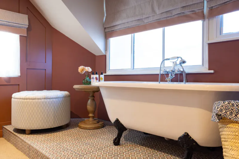 Elegant clawfoot bathtub in luxurious red-walled bathroom with ottoman, flowers, and window light.