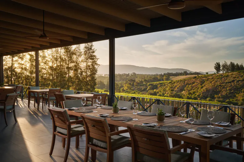 Elegant wooden dining tables set on a covered terrace at Brookdale Wine Estate, overlooking sunset hills and vineyards in Paarl.