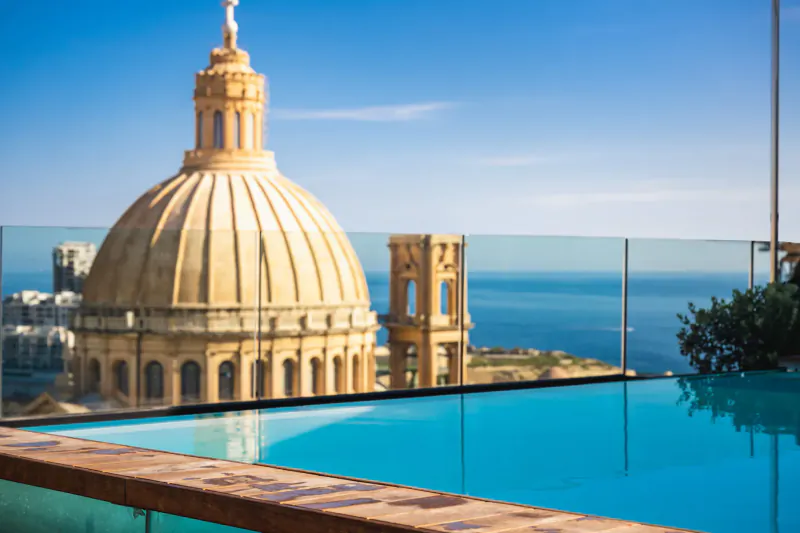 Valletta's golden domed cathedral and bell tower viewed from rooftop infinity pool with sea beyond, blue sky.