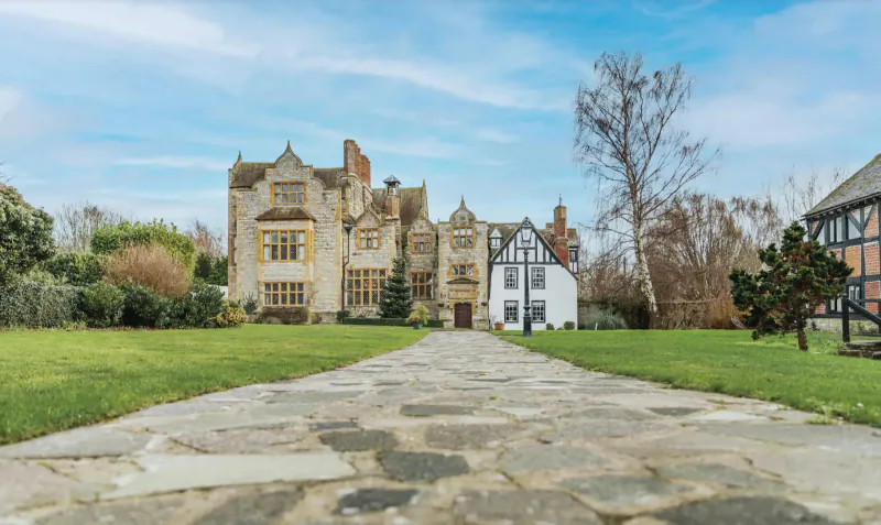 Karma Salford Hall, a Tudor-style mansion with white cottage, fronted by stone path in lush gardens under blue sky.