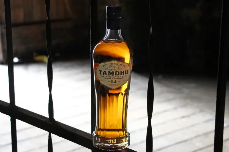 Tamdhu 12 whisky bottle with amber liquid, on wooden surface behind black metal bars.