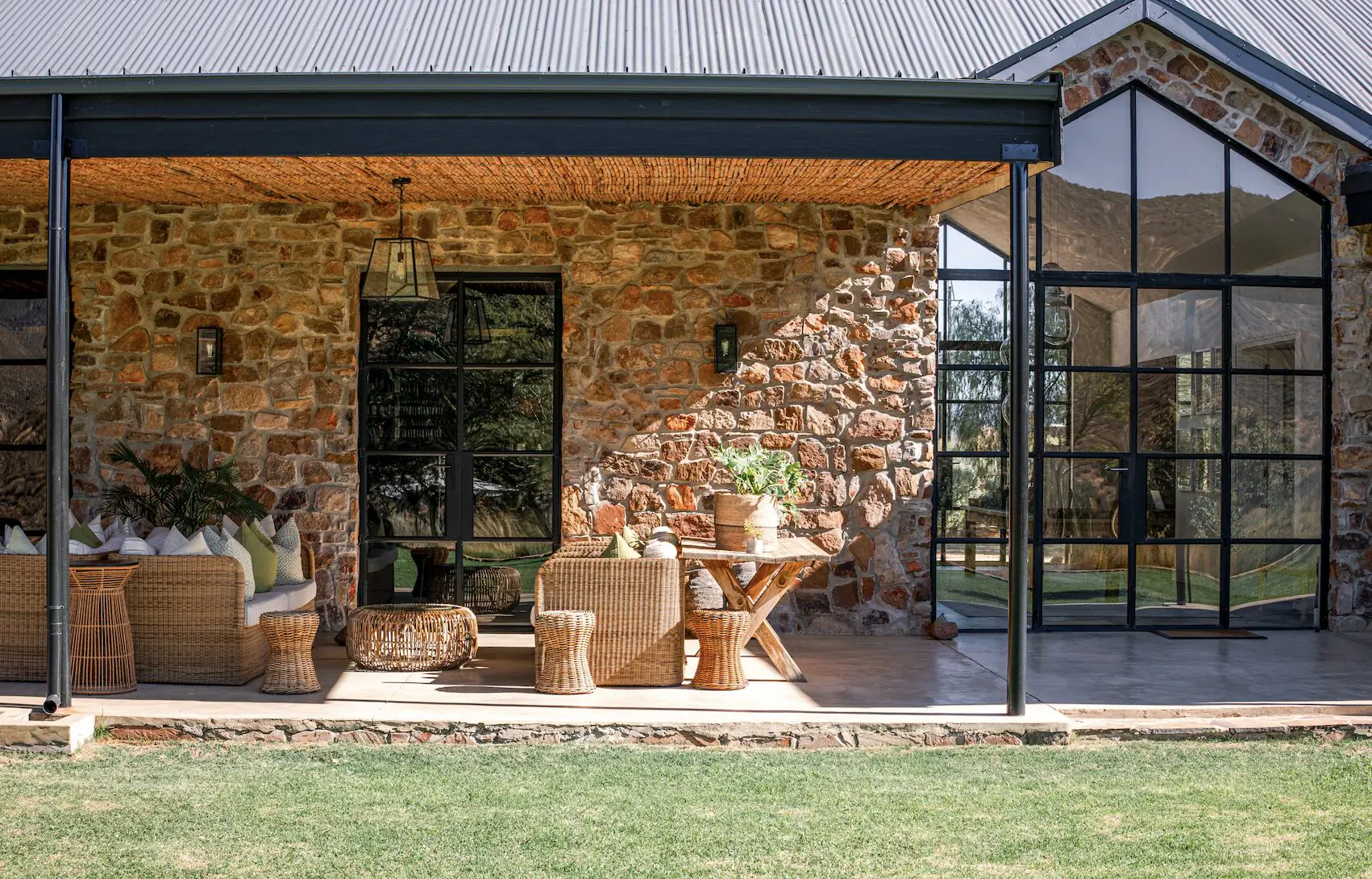 Rustic stone house veranda with black-framed glass doors, wicker furniture, plants, and table on green lawn