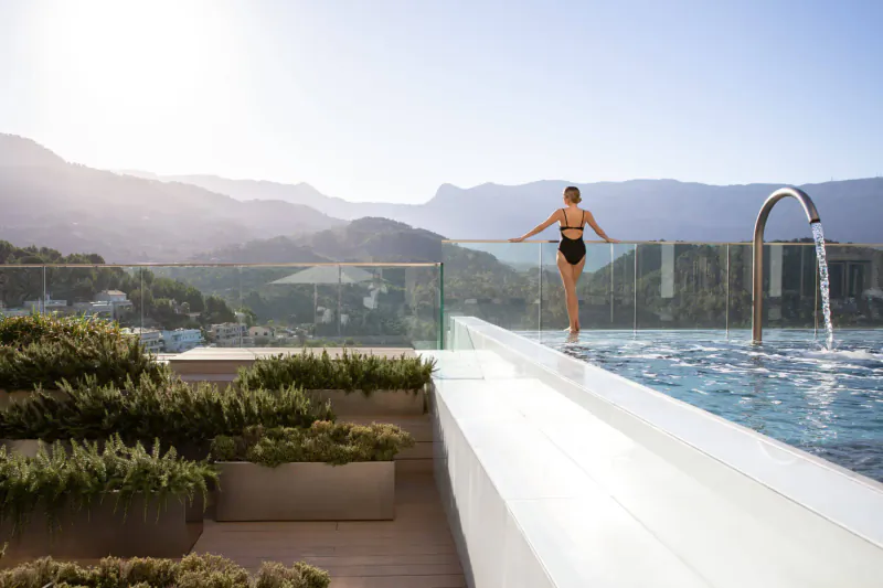 Woman in black bikini stands at infinity pool edge on hotel rooftop, mountains and sea view, Mallorca.