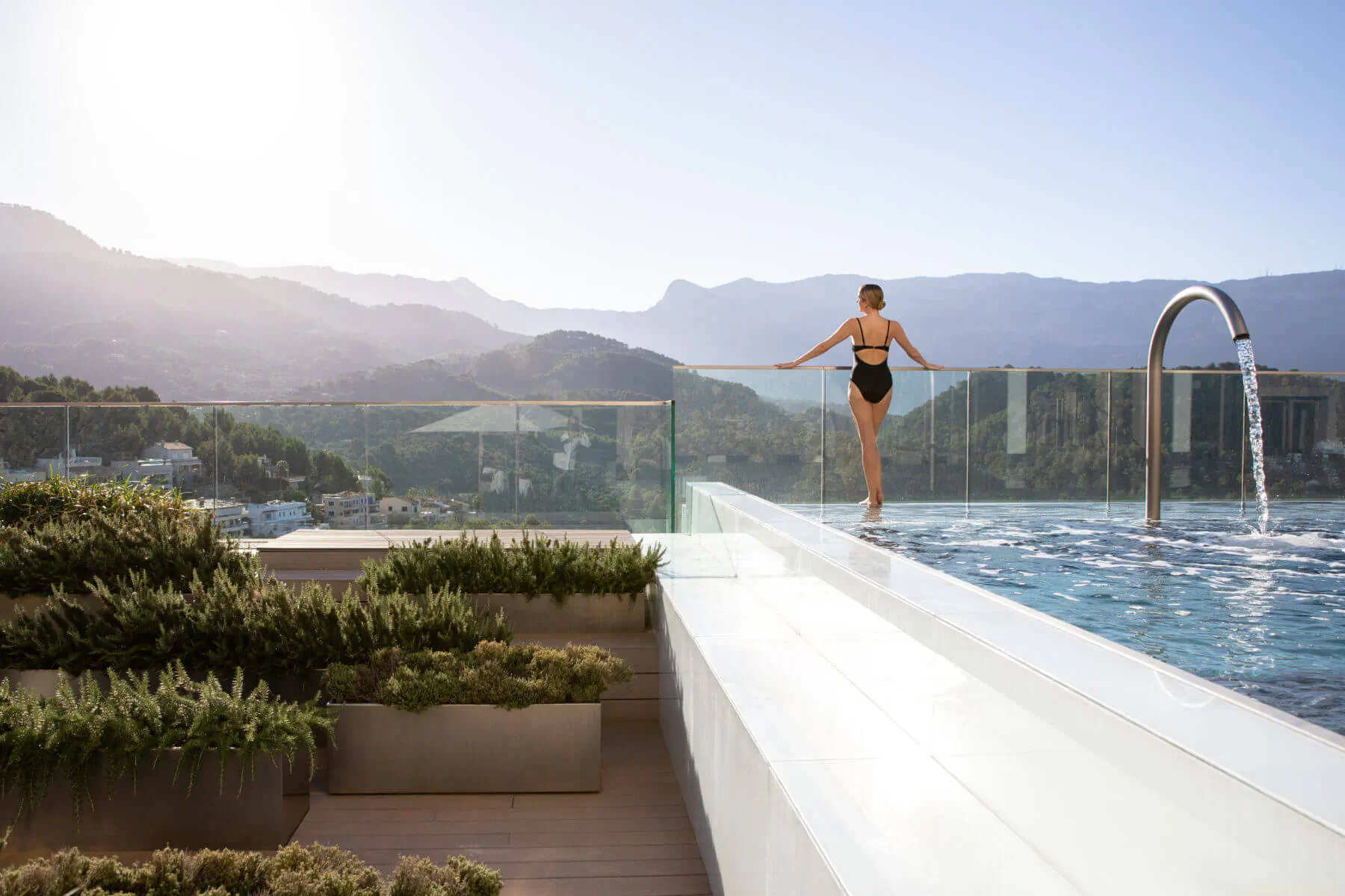Woman in black bikini stands at infinity pool edge on hotel rooftop, mountains and sea view, Mallorca.