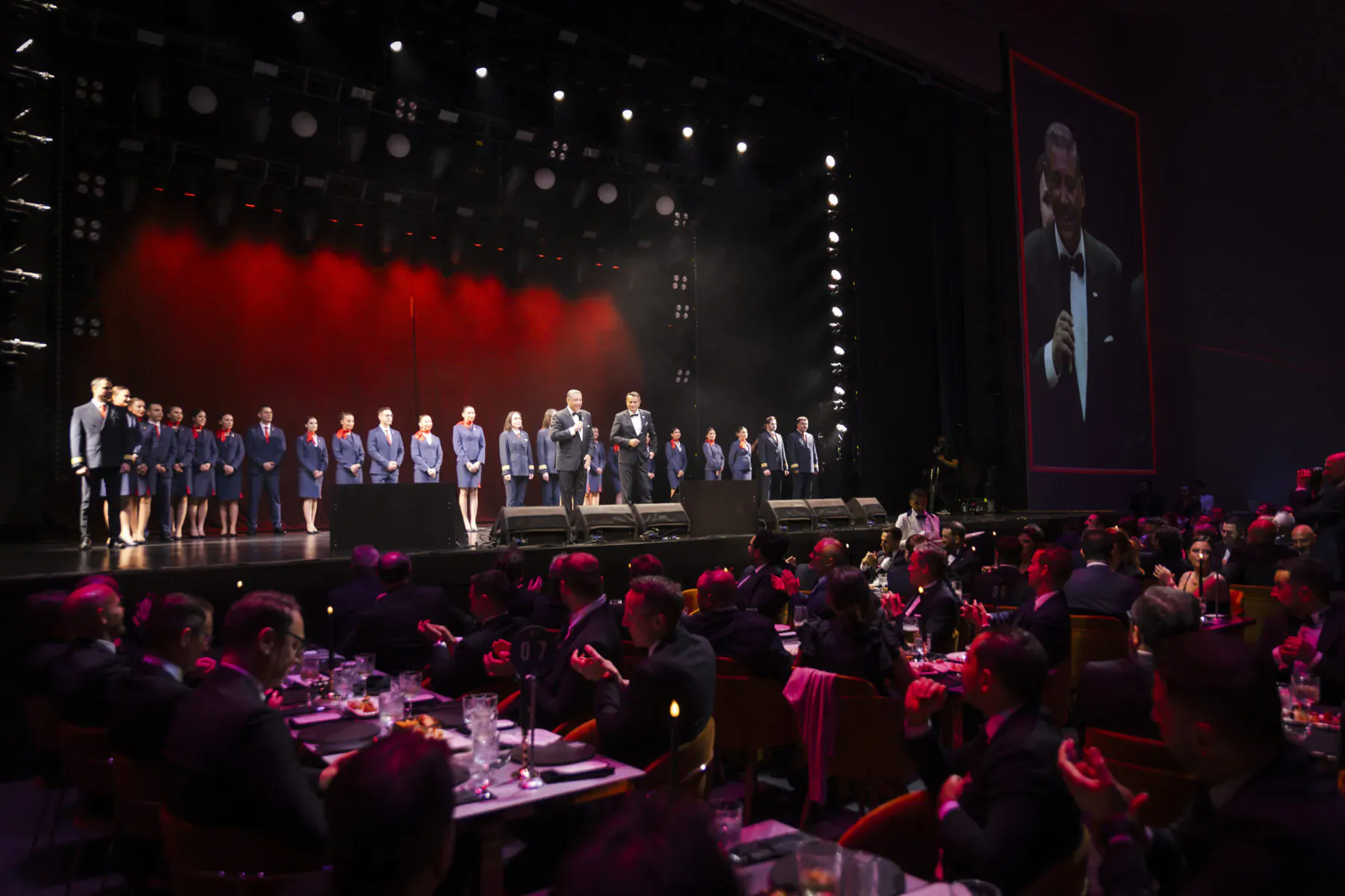 Choir in blue uniforms performing on red-lit stage at Corendon Airlines anniversary gala with formal audience at candlelit tables