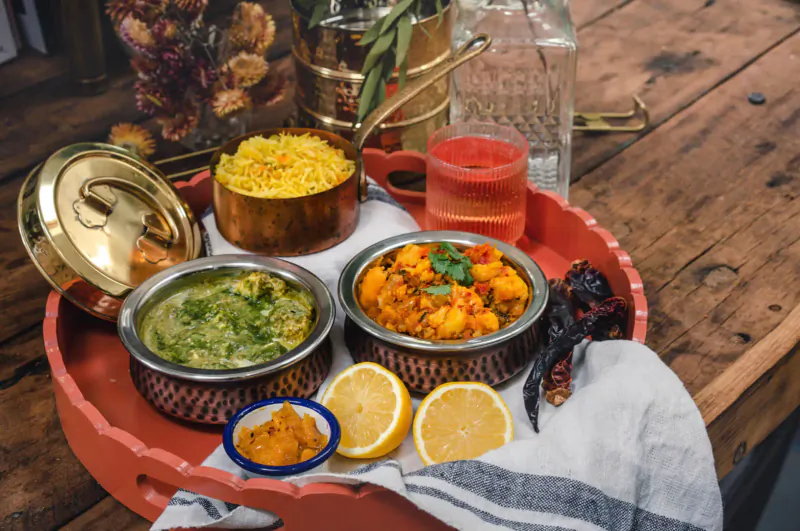 Indian dishes on red tray: yellow rice, green curry, chickpea masala, chutney, lemon wedges, red chilies, on wooden table