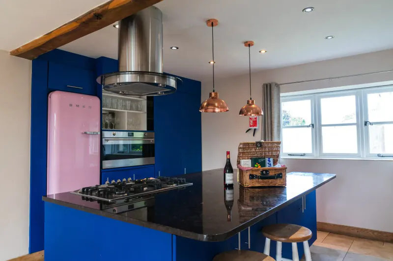 Modern Cotswolds kitchen with pink Smeg fridge, blue cabinets, stainless hood, copper pendants, wine bottle on black island.