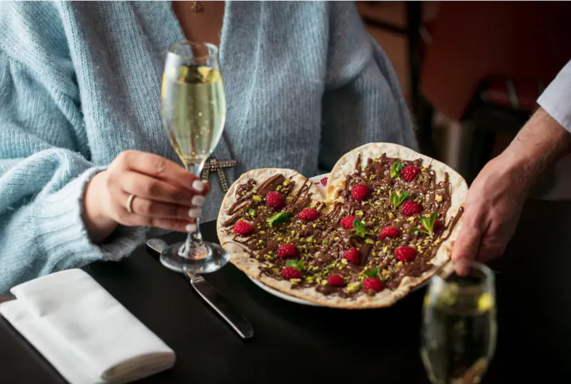 Woman and chef holding heart-shaped dessert pizza topped with raspberries and pistachios, champagne glasses on table.