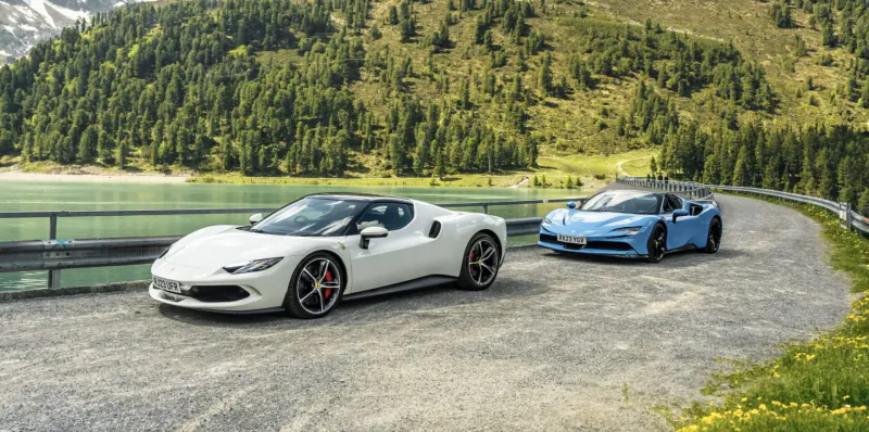 White and blue Ferrari SF90s parked on a mountain road by alpine lake amid forests and peaks.