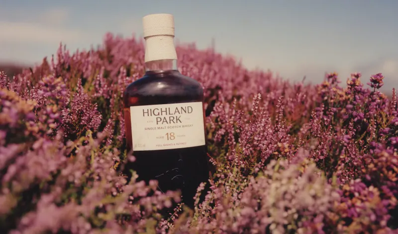 Highland Park whisky bottle in purple heather field under blue sky, label reads 'Highland Park 18'.