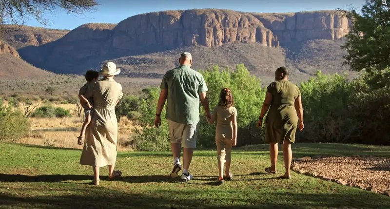 Family in safari attire walks hand-in-hand on lawn with African mountains and trees at Marataba Game Lodges.