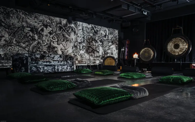 Dimly lit Mandrake hotel gallery with large gongs, green yoga mats and pillows on floor, abstract black-and-white wall mural.