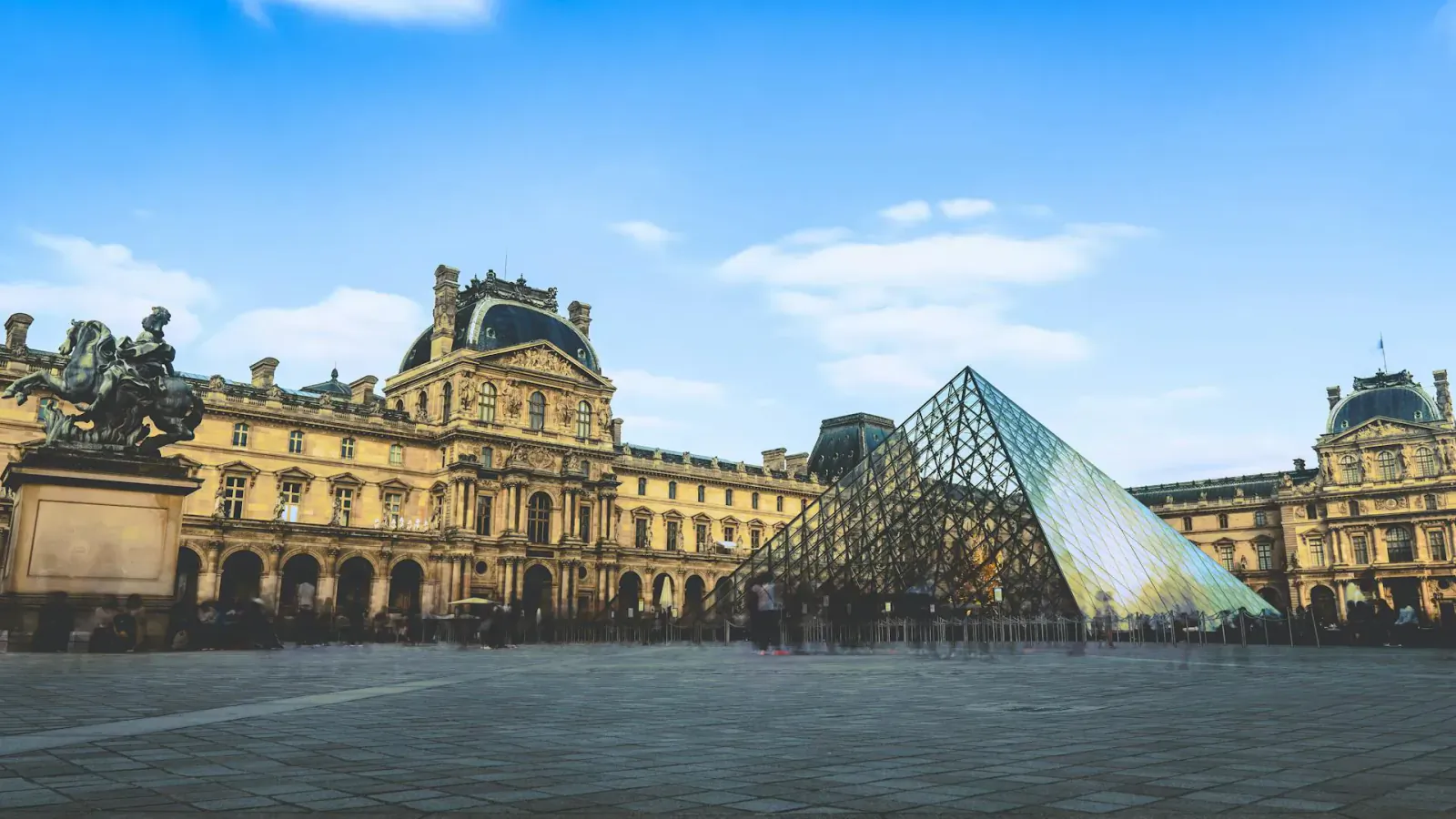 Louvre Pyramid in the Palais du Louvre courtyard, Paris, with statues and people under blue sky.