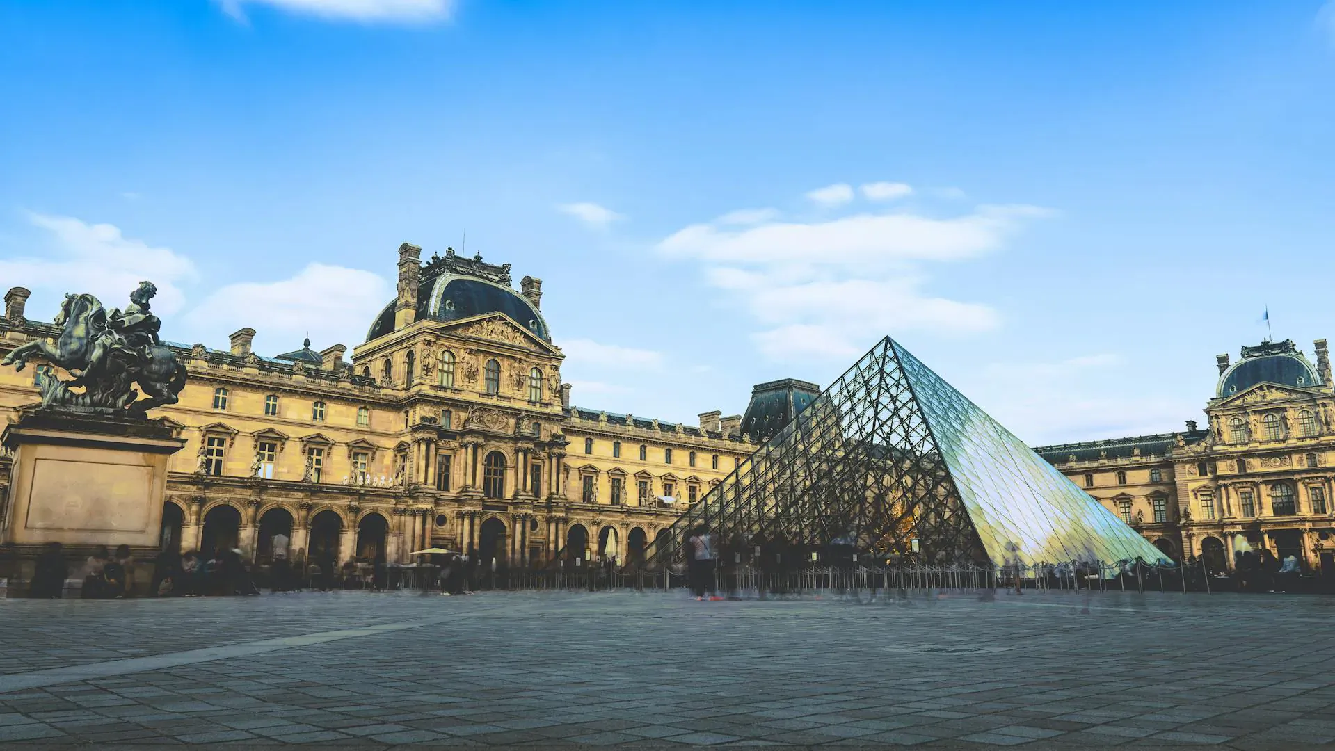 Louvre Pyramid in the Palais du Louvre courtyard, Paris, with statues and people under blue sky.