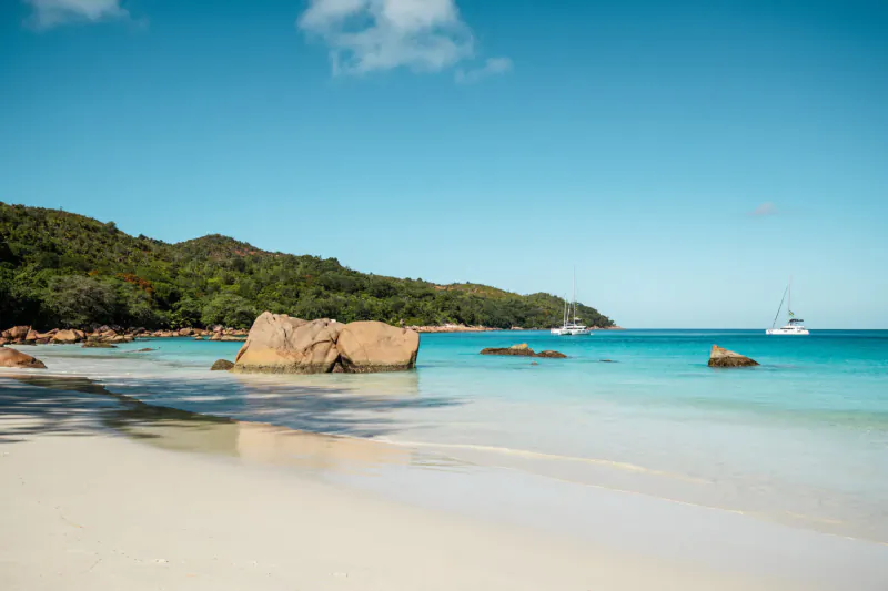 Praslin, Seychelles beach with large granite boulders, turquoise ocean, sailboats, lush green hills under blue sky