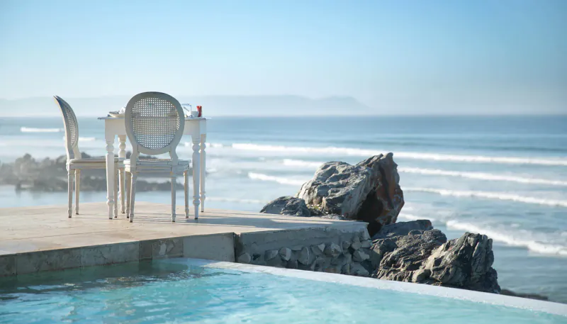 White chairs and table on infinity pool deck overlooking ocean waves and rocks in Hermanus