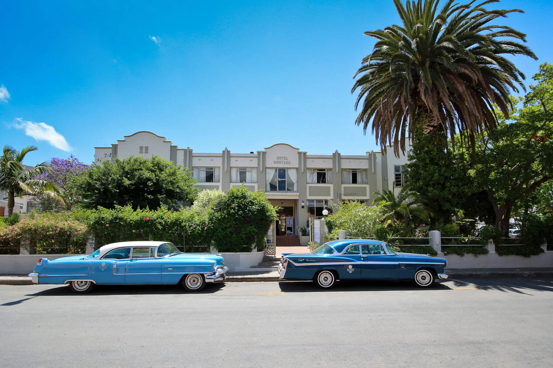 Montagu Country Hotel facade with two blue classic cars parked in front, palm trees, lush gardens, blue sky.