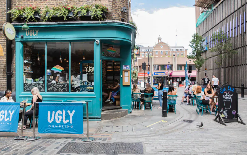 Ugly Mugs café exterior in Carnaby London with blue facade, outdoor seating, patrons, and 'UGLY' sign on cobblestone street.