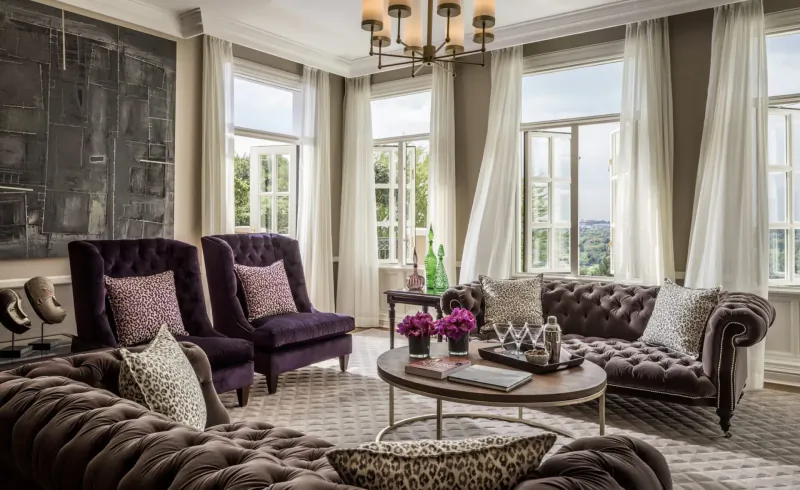 Elegant hotel living room at Four Seasons Westcliff Johannesburg with purple velvet armchairs, tufted sofa, chandelier, flowers, and city view.