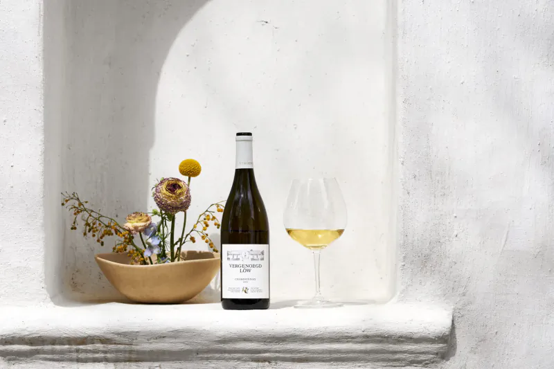 Vergenoegd Löw Wine Estate bottle of white wine and wine glass beside dried yellow flowers in terracotta bowl on sunlit white ledge