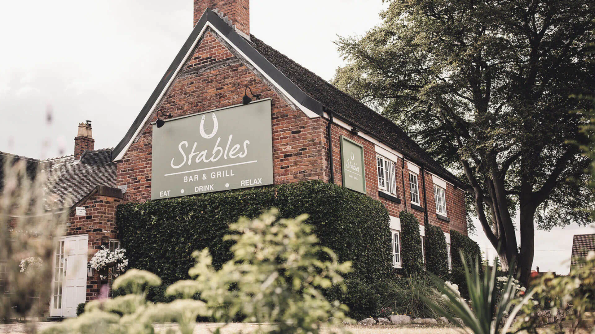 Stables Bar & Grill sign on brick building at The Manor House Hotel & Spa Cheshire, with trees and hedges.