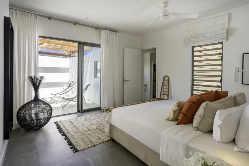 Bright beachfront bedroom in Villa Ruby, Mauritius, with white bed, orange pillows, rattan chair, large vase, and balcony view.