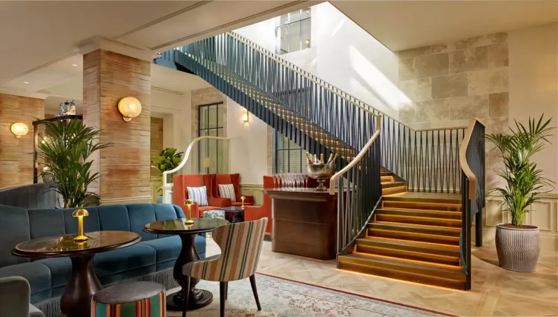 Elegant hotel lobby with curved dark blue staircase, plush velvet seating, plants, and warm lighting