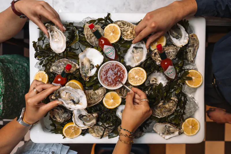 Overhead view of fresh oysters on seaweed with lemon wedges, Tabasco bottles, and cocktail sauce on a white platter, hands shucking them.