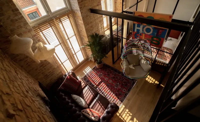 Top-down view of cozy loft suite with red sofa, swing bed, plants, exposed brick, large windows, and 'HOTEL' wall art.