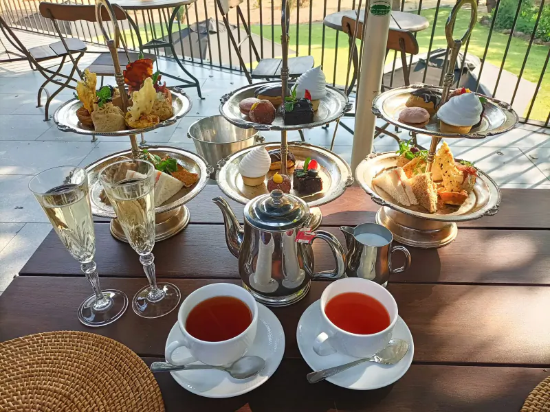 Elegant afternoon tea on outdoor wooden table at luxury hotel: tiered stands of scones, cakes, sandwiches, teapot, tea cups, champagne flutes.