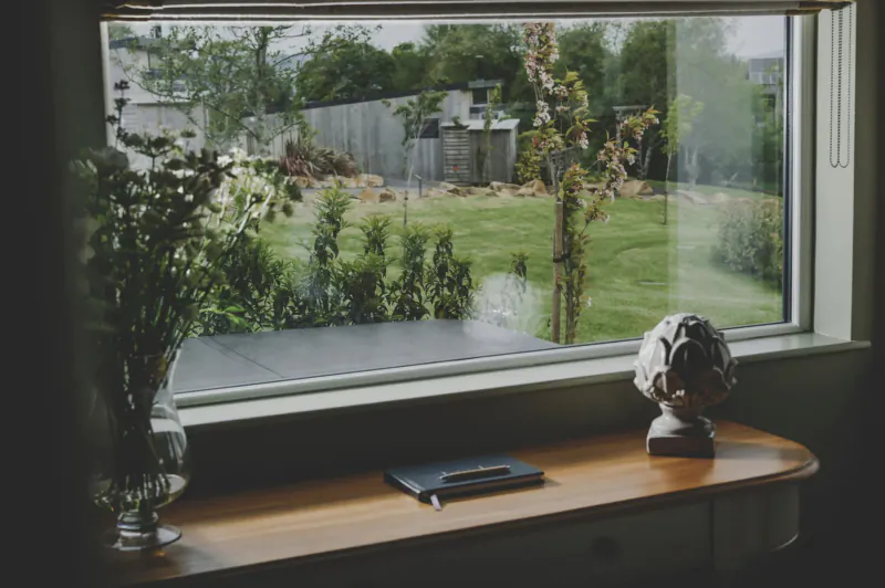 Window view of lush garden with trees, hedges, and lawn at Everything Retreat; wooden desk with notebook, vase of flowers, stone bust.