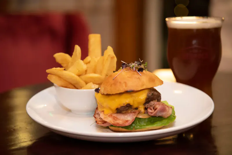 Bacon cheeseburger with microgreens and fries on white plate, beside beer pint on wooden table