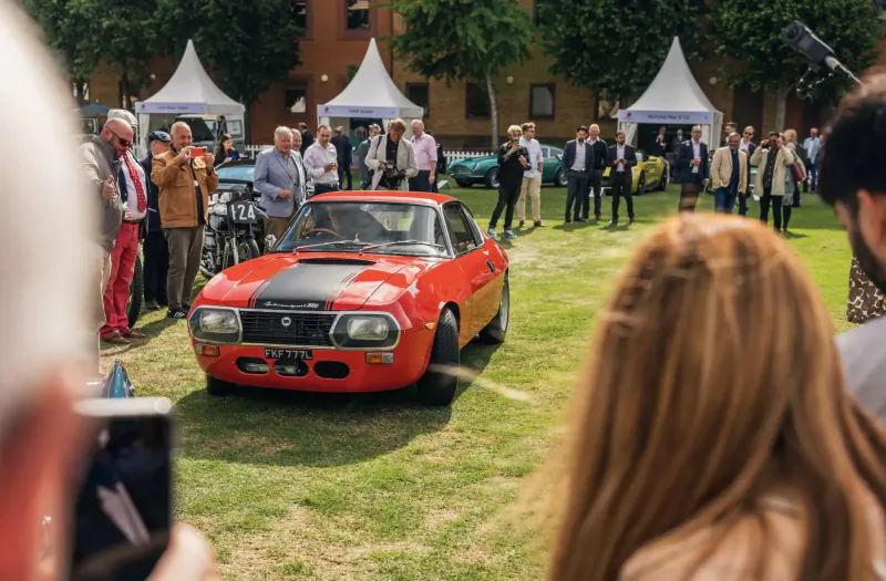 Red Mazda Cosmo sports car with black hood scoop displayed at London Concours 2024, surrounded by crowd on grassy field with white tents.